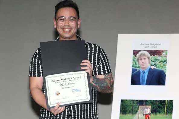 A smiling person holds an open certificate folder displaying a “Shaina Strelzman Award.” Beside them is a poster with two photographs and the name Andrew Helgason.