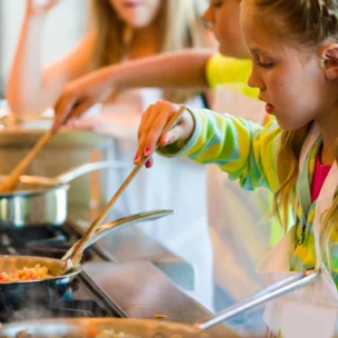 A young girl stirs food in a pan on a stove, wearing a striped shirt and apron, with other children cooking in the background.
