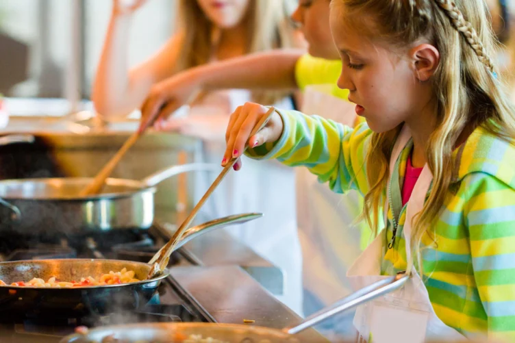 A young girl stirs food in a pan on a stove, wearing a striped shirt and apron, with other children cooking in the background.