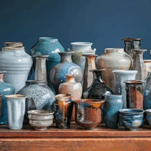 A collection of assorted ceramic vases, bowls, and cups in various shapes, sizes, and earthy tones, arranged on a wooden surface against a dark blue background.