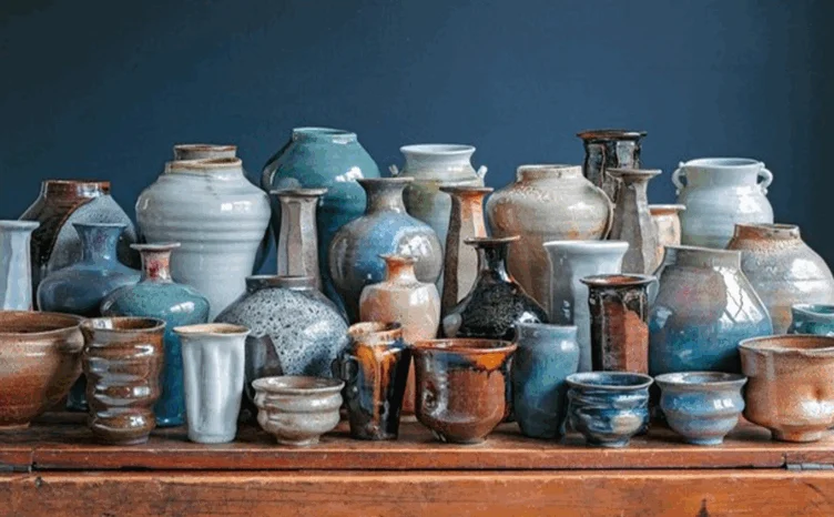 A collection of assorted ceramic vases, bowls, and cups in various shapes, sizes, and earthy tones, arranged on a wooden surface against a dark blue background.
