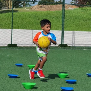 A young boy holding a yellow ball runs through green and blue cones on an outdoor sports field.