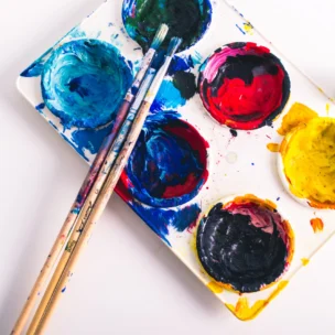 A white palette with colorful paint, three paintbrushes, an open yellow paint tube, and a small yellow paint container on a white surface.