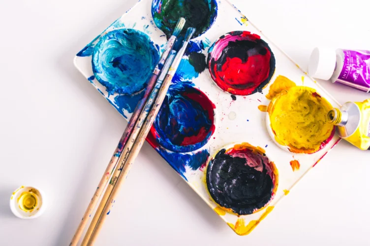 A white palette with colorful paint, three paintbrushes, an open yellow paint tube, and a small yellow paint container on a white surface.