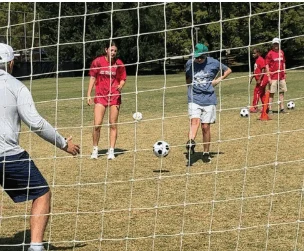 A group of people practice soccer on a grass field, with one person preparing to kick a ball toward a goal while others observe.