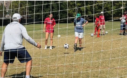 A group of people practice soccer on a grass field, with one person preparing to kick a ball toward a goal while others observe.