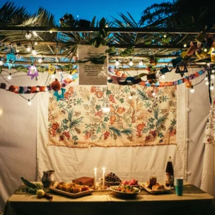 A decorated sukkah with hanging lights, paper chains, and a table set with candles, fruits, bread, and wine, prepared for a festive meal.