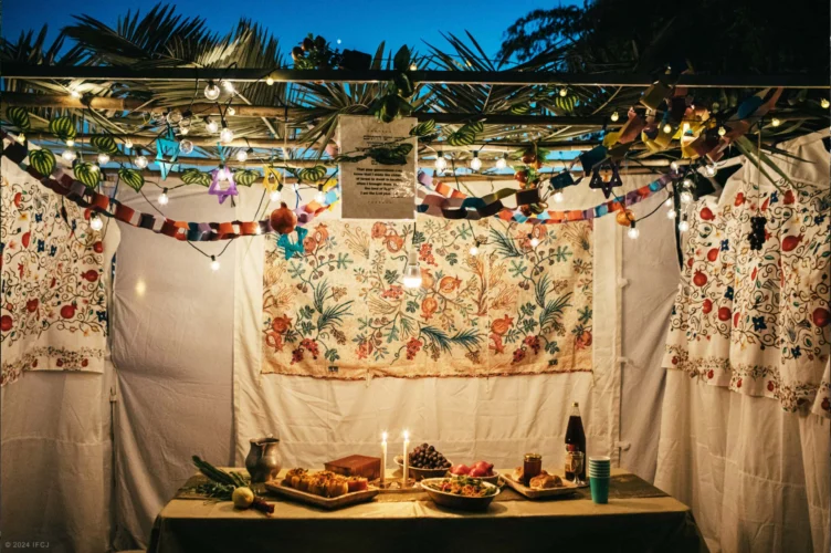 A decorated sukkah with hanging lights, paper chains, and a table set with candles, fruits, bread, and wine, prepared for a festive meal.