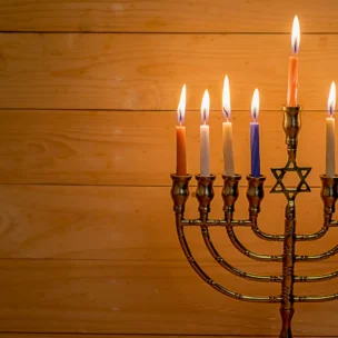 A lit nine-branched menorah with candles of various colors stands against a wooden background.