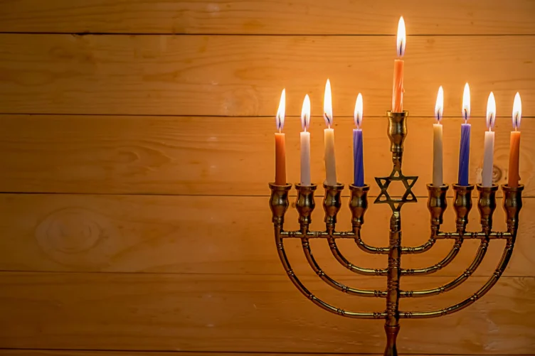 A lit nine-branched menorah with candles of various colors stands against a wooden background.