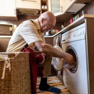 An older man kneels in a kitchen, loading clothes into a front-loading washing machine beside a wicker laundry basket.