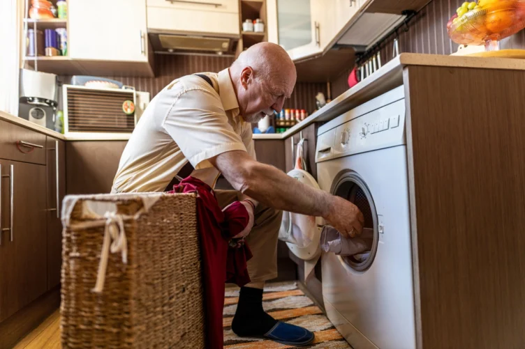 An older man kneels in a kitchen, loading clothes into a front-loading washing machine beside a wicker laundry basket.