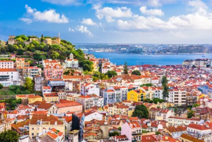 A panoramic view of Lisbon with colorful buildings, red rooftops, São Jorge Castle on a hill, and the Tagus River in the background under a partly cloudy sky.