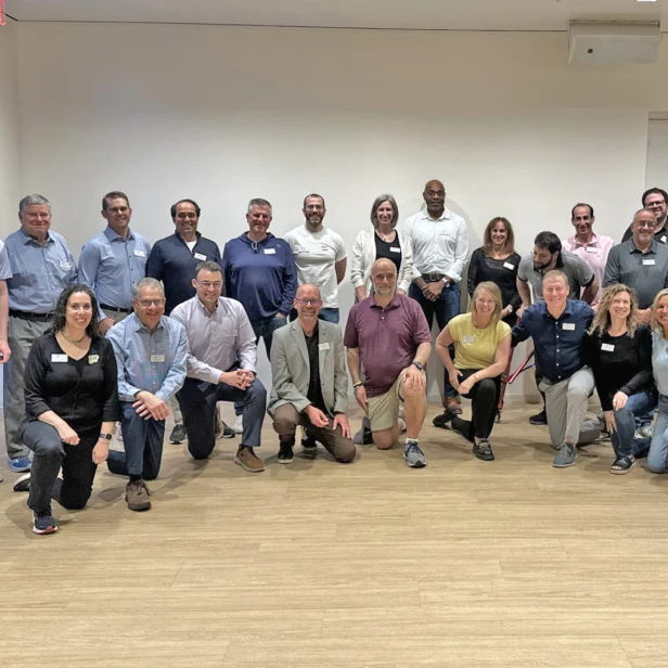 A group of about 30 adults, both men and women, pose together indoors in two rows, some standing and some kneeling, in a brightly lit room with wooden floors.