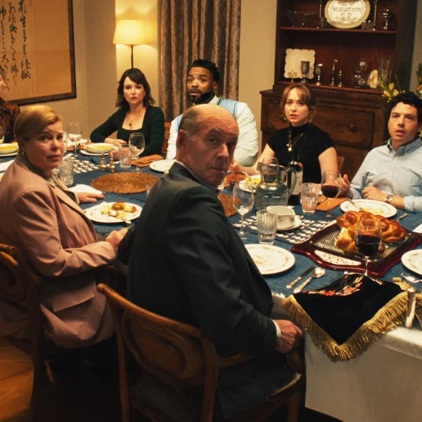 A group of people sit around a dinner table set with food, all turning to look toward the camera with surprised or serious expressions.