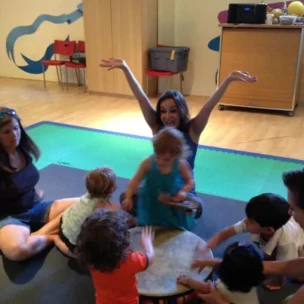 Adults and children sit on the floor in a circle, playing a large drum together, while one woman raises her arms and smiles energetically.