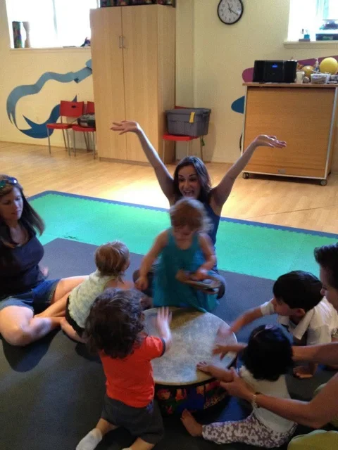 Adults and children sit on the floor in a circle, playing a large drum together, while one woman raises her arms and smiles energetically.