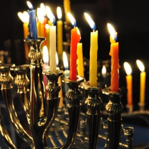 A row of lit candles of various colors in a silver menorah, with some flames burning brightly against a dark background.