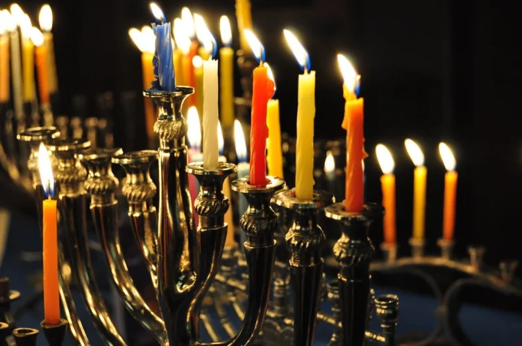 A row of lit candles of various colors in a silver menorah, with some flames burning brightly against a dark background.