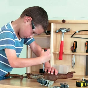 A young boy wearing safety glasses uses a screwdriver on a metal object at a workbench with various hand tools and a wooden tool organizer.