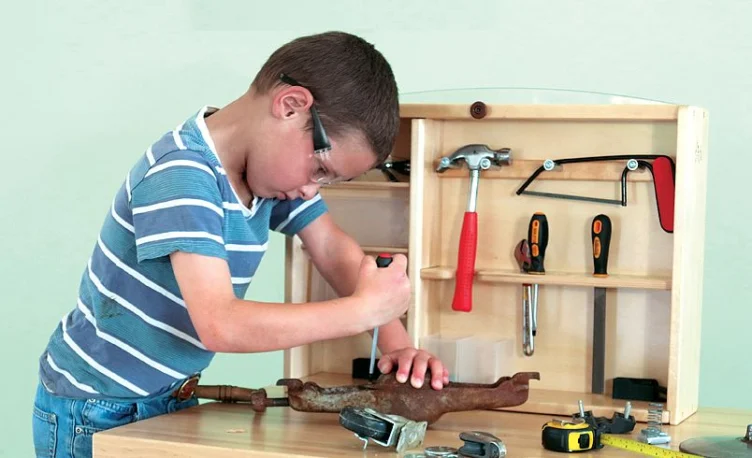 A young boy wearing safety glasses uses a screwdriver on a metal object at a workbench with various hand tools and a wooden tool organizer.