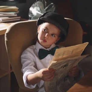 A young child dressed in a white shirt, bow tie, and cap sits in an office chair, holding a newspaper and looking to the side with a curious expression.