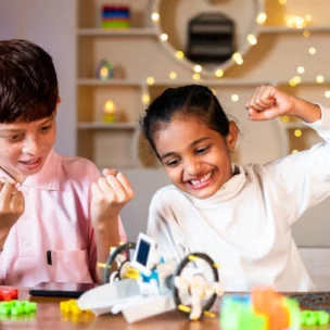 Two children sitting at a table cheerfully celebrating while playing with colorful building blocks and a small robot.