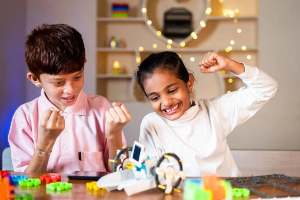 Two children sitting at a table cheerfully celebrating while playing with colorful building blocks and a small robot.