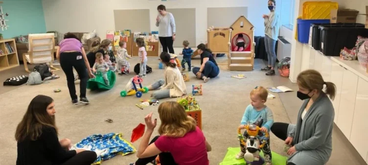 Several adults and young children interact and play with toys in a spacious indoor playroom, some adults are seated, and a few are wearing face masks.