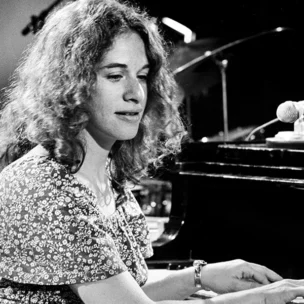 A woman with curly hair plays a Steinway & Sons grand piano on stage, with a microphone nearby.
