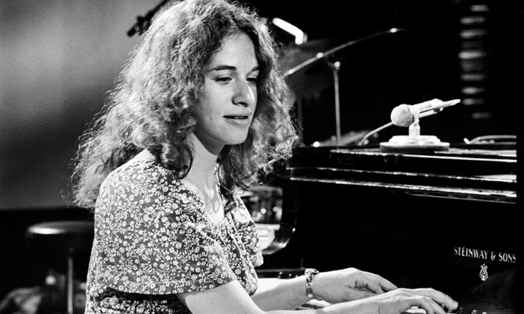 A woman with curly hair plays a Steinway & Sons grand piano on stage, with a microphone nearby.