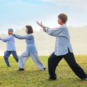 Three people practice tai chi outdoors on grass near a body of water, each holding a pose and wearing loose, comfortable clothing.