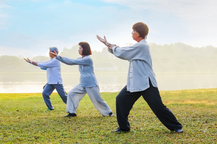 Three people practice tai chi outdoors on grass near a body of water, each holding a pose and wearing loose, comfortable clothing.