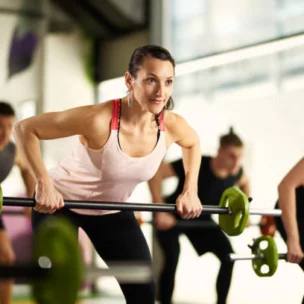 A group of people exercise indoors, lifting barbells in a fitness class; a woman in a pink tank top is in the foreground, focused on her workout.