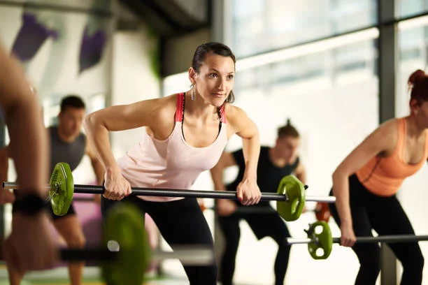 A group of people exercise indoors, lifting barbells in a fitness class; a woman in a pink tank top is in the foreground, focused on her workout.