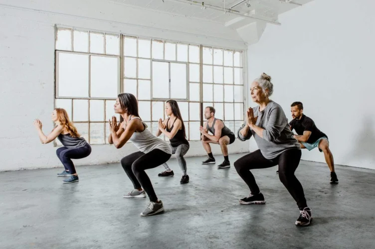A group of people in athletic wear performing squats in a bright, spacious room with large windows.