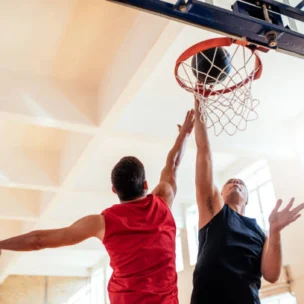 Two men playing basketball indoors; one in a red jersey attempts a layup while the other in a black jersey tries to block the shot near the hoop.