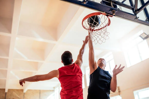 Two men playing basketball indoors; one in a red jersey attempts a layup while the other in a black jersey tries to block the shot near the hoop.