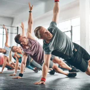 A group of people in athletic clothing perform side plank exercises on the floor in a gym with large windows.