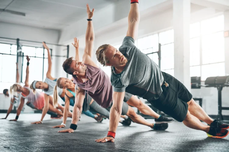 A group of people in athletic clothing perform side plank exercises on the floor in a gym with large windows.