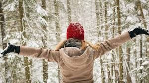 A person in a tan coat and red hat stands in a snowy forest with arms outstretched, facing away from the camera.
