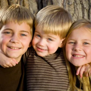 Three smiling children, two boys and one girl, pose closely together in front of a large tree trunk outdoors.