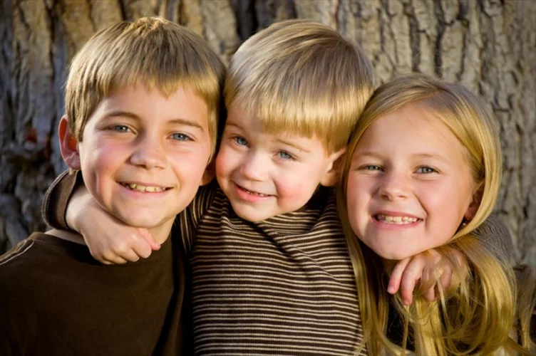 Three smiling children, two boys and one girl, pose closely together in front of a large tree trunk outdoors.