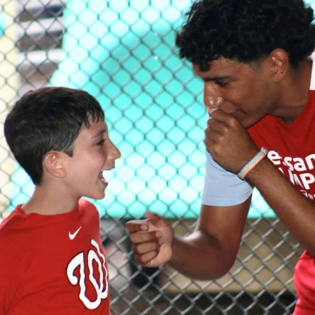 A smiling boy and an older youth in red shirts face each other, appearing to be in an animated conversation near a chain-link fence.