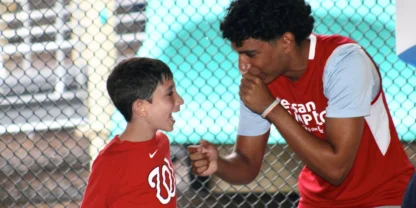 A young boy and a teen, both in red shirts, enthusiastically interact in front of a chain-link fence.
