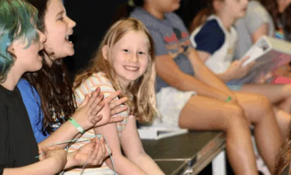 A group of children sit in a row on a bench, some clapping and laughing, while one child in the center looks at the camera and smiles.