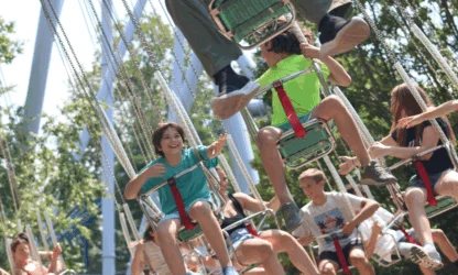 Children and teenagers ride on outdoor swing carousel chairs, smiling and enjoying a sunny day at an amusement park. Trees and metal ride structure are visible in the background.