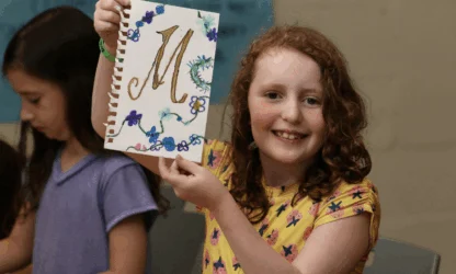 A smiling girl holds up a drawing with a large gold letter "M" surrounded by flowers, while another girl sits beside her.