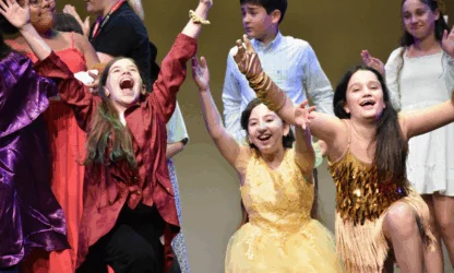Three children in colorful costumes smile and raise their arms enthusiastically on stage, surrounded by other kids in various outfits.