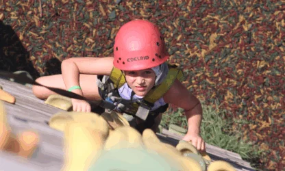 A person wearing a red helmet and safety harness is climbing an outdoor artificial rock wall, looking upward.